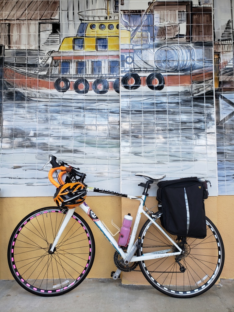 a white road bicycle with black panniers, orange handlebars, orange helmet, pink and white rim stickers, and pink water bottle, in front of a mural of a harbor.