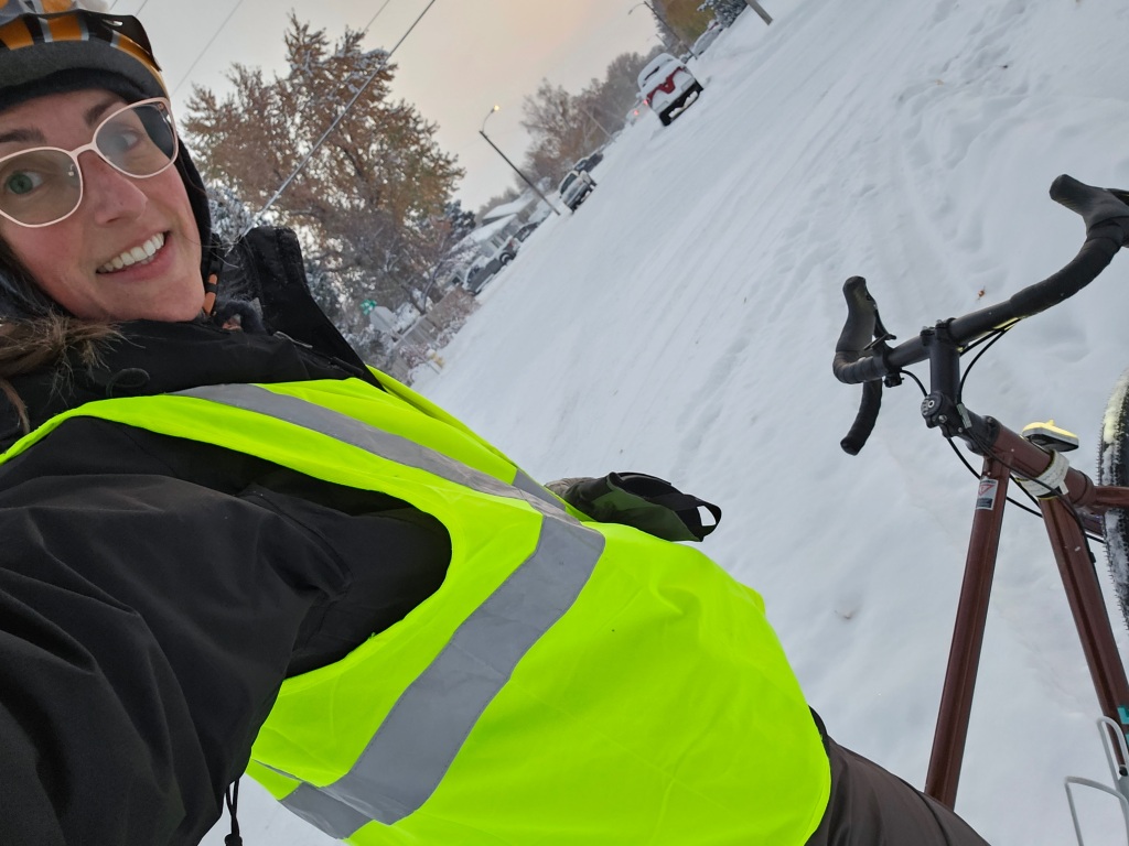 a young white female with pink glasses, orange bicycle helmet, and yellow reflective vest standing over her brown bicycle on a very snowy road during the morning commute home