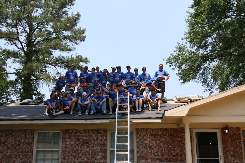 34 young adults in blue t-shirts standing & sitting in a group on top of a roof on a construction site