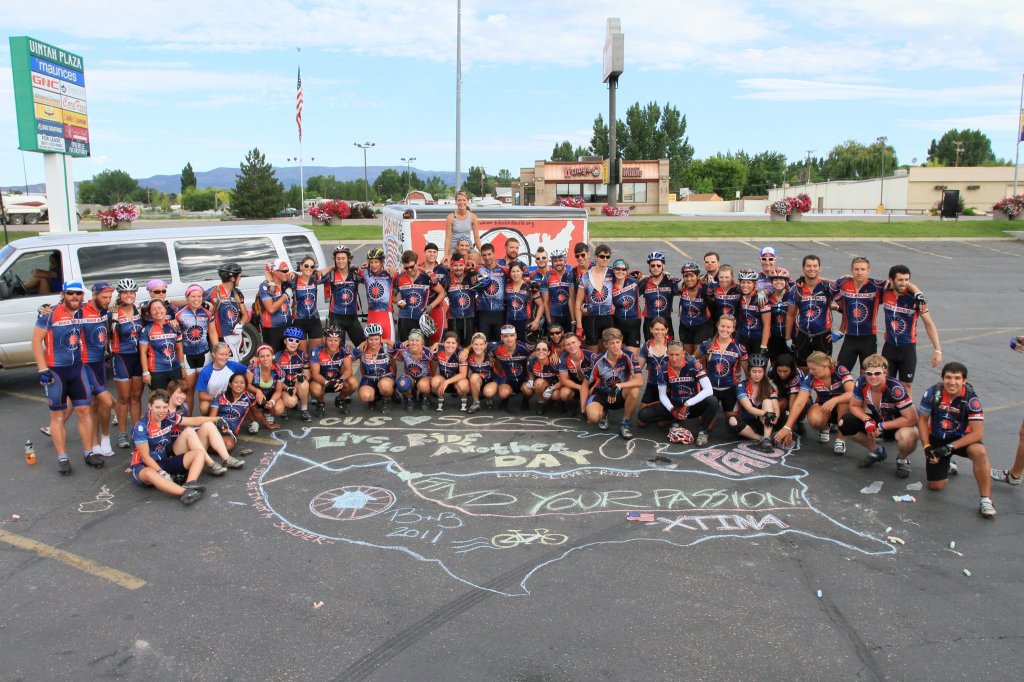 70 cyclists in navy jerseys and black shorts in a parking lot posing together around a chalk drawing of the United States with each team's cross-country routes drawn out, including memorial notes for deceased riders