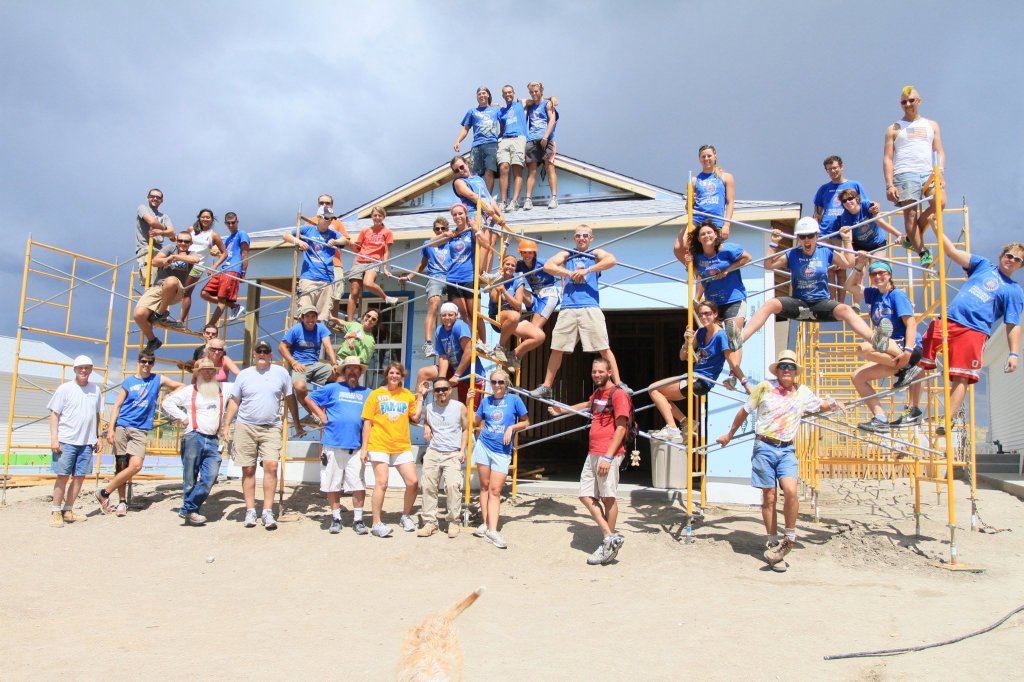 40 adults of various ages, genders, races, and sizes standing on and in front of scaffolding on the construction site of a mostly finished Habitat for Humanity  house