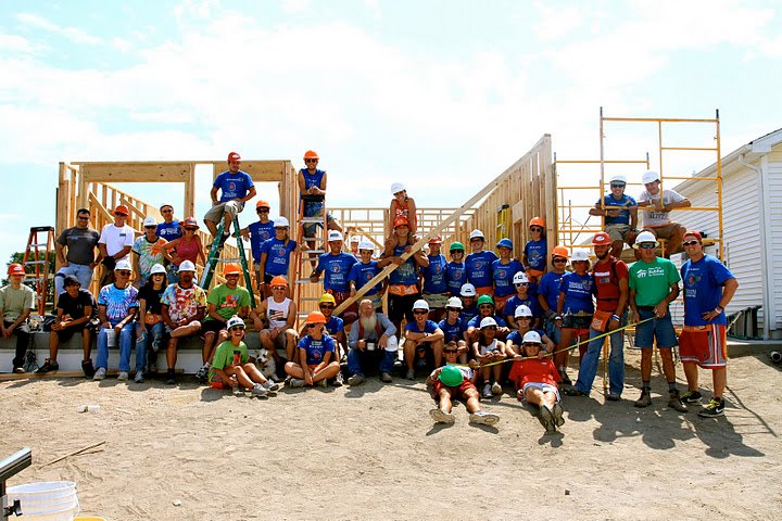40 adults wearing hard hats posing in front of an open framed house on a construction site 